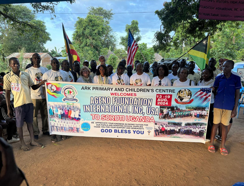A group of people, including adults and children, stands outdoors holding a large banner. The banner welcomes the Ageno Foundation International Inc., USA, to Soroti, Uganda. Several flags and text are visible on the banner. Trees and a building are in the background.