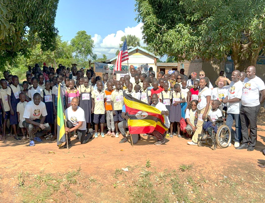 A large group of children and adults pose outdoors in front of a building. They hold flags, including Ugandan and American flags. Some wear matching shirts, and one person is in a wheelchair. Trees and a blue sky are in the background.