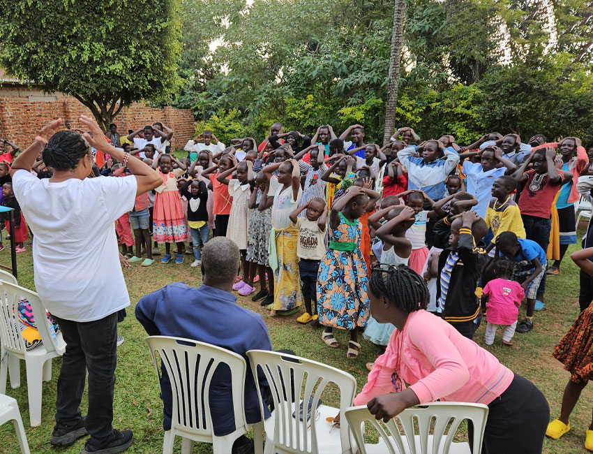 A group of children and adults gathered outdoors, following a person leading them in a hand movement exercise. Some people are seated on white plastic chairs. Trees and a brick wall are visible in the background.