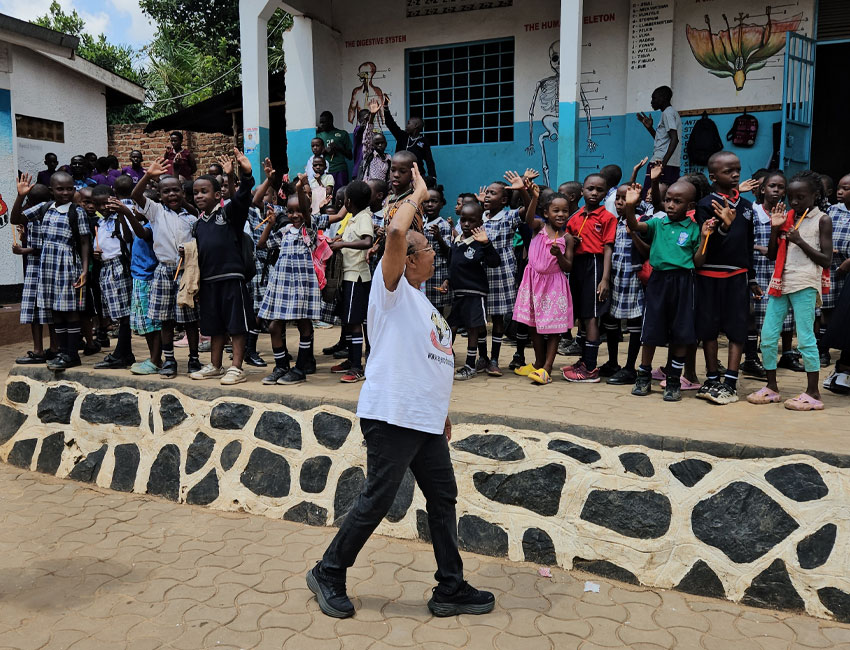 A person leads a group of children standing on a platform at a school. The children are wearing uniforms and some casual clothes, waving their hands. The background has educational posters and a mural of a human skeleton.