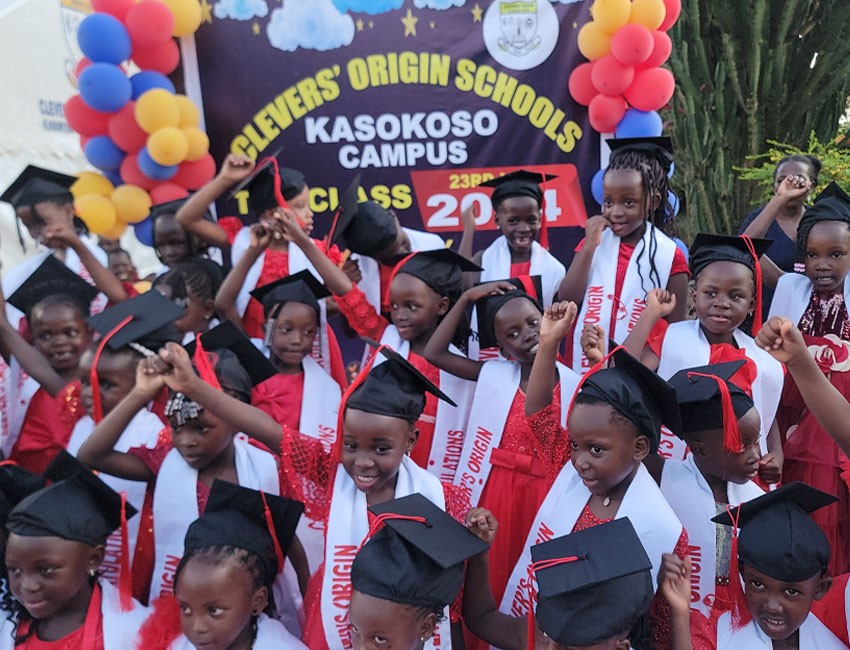 A group of children wearing graduation caps and sashes celebrate at a graduation event. They stand in front of a colorful balloon arch. A banner reads "Clever's Origin Schools, Kasokoso Campus, 1st Class 2024.