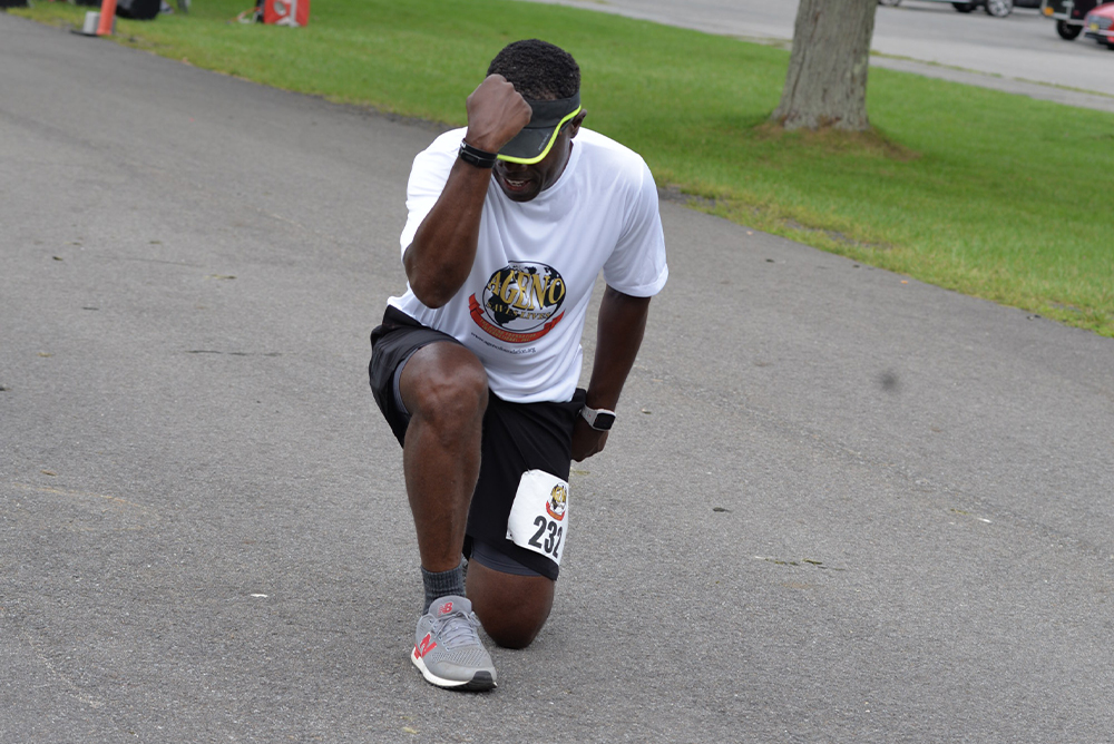 A person wearing a white t-shirt, black shorts, and running shoes is kneeling on one knee on a road, touching their forehead with their hand. They have a race bib on their left leg, indicating they are participating in a charity run. A grassy area with a tree is visible in the background.