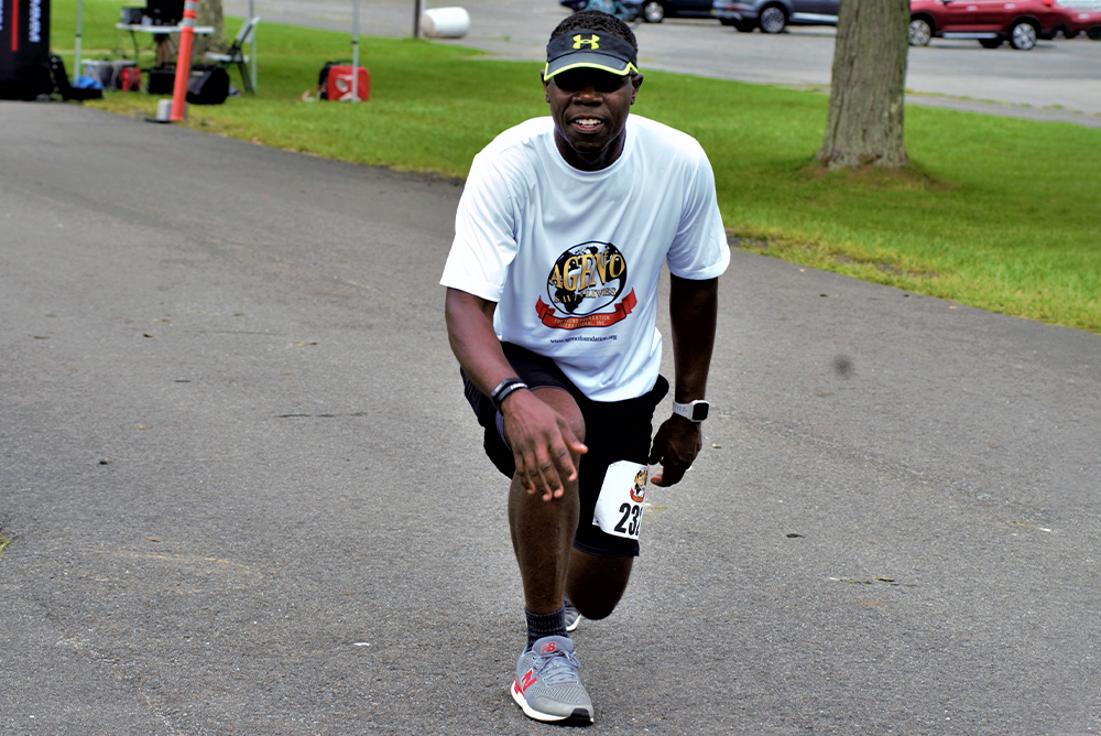 A man wearing a white t-shirt, black shorts, white and gray running shoes, and a black cap is crouching on a paved path in a park. He has a race number pinned to his shorts and is smiling at the camera. Grass, trees, and parked cars are visible in the background as he prepares for the charity run.