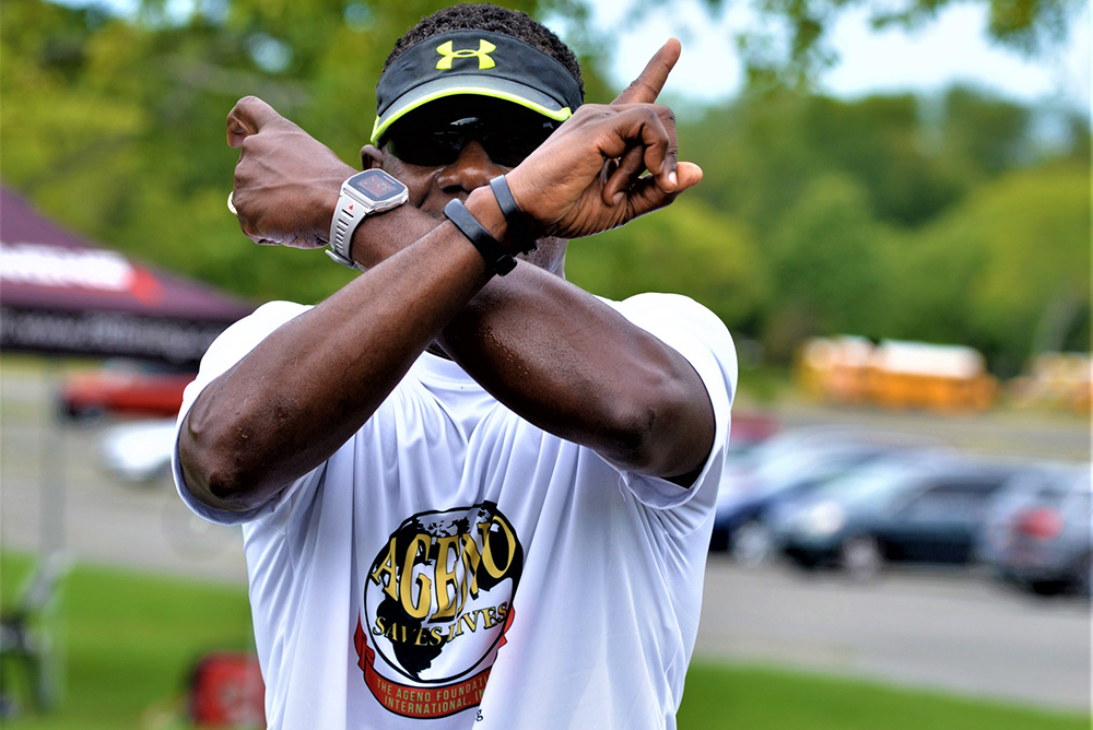 A person wearing a white T-shirt with a colorful design promoting charity and a black visor is standing outdoors with their arms crossed in an "X" in front of their face. The background shows trees, vehicles, and a partly cloudy sky, reflecting the spirit of a water donation event.