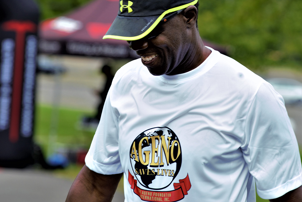A person wearing sunglasses and a hat smiles while looking down. Dressed in a white T-shirt with a colorful logo that reads "AGEN Foundation Saves Lives," they seem to be outdoors, possibly at a charity event or gathering, with blurred people and tents in the background.