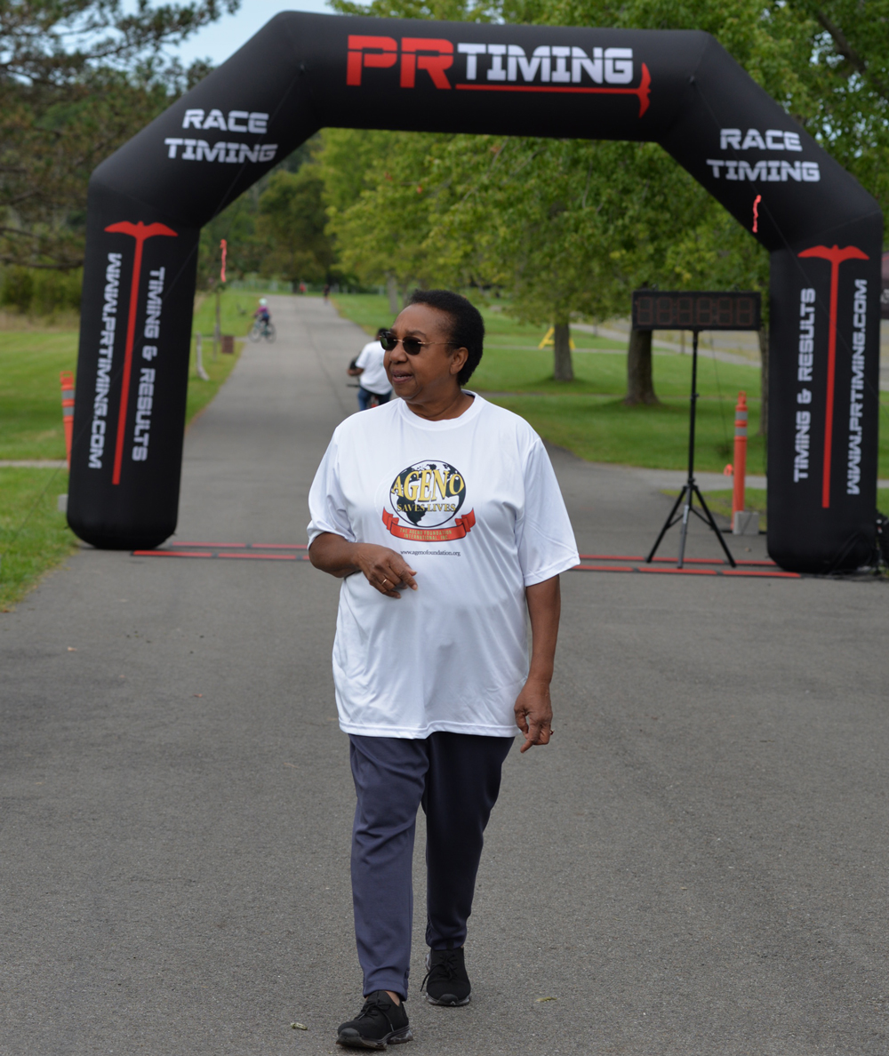 A person wearing a white t-shirt, dark pants, and sunglasses walks past a black race timing archway on a paved road. The person has their hands in their pockets and looks to the side. Trees and grass line the road in the background with a few other people in the distance, near water stations for charity donations.