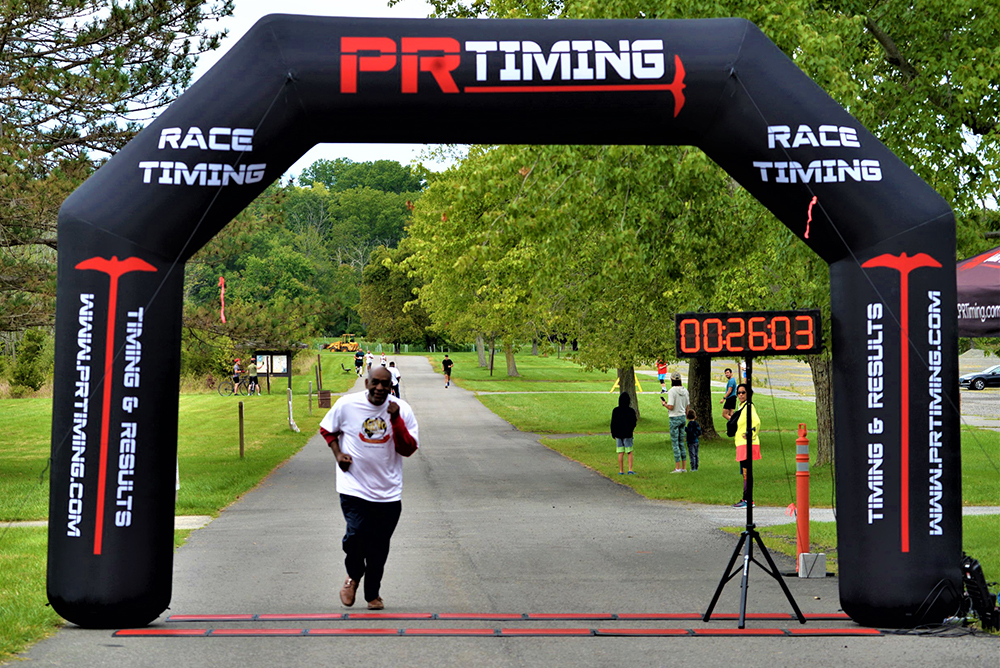 A runner wearing a white shirt crosses the finish line under an inflatable arch that reads "PR Timing." A digital clock displays a time of 02:26:03. The scene, part of a charity race, features trees and spectators in the background.