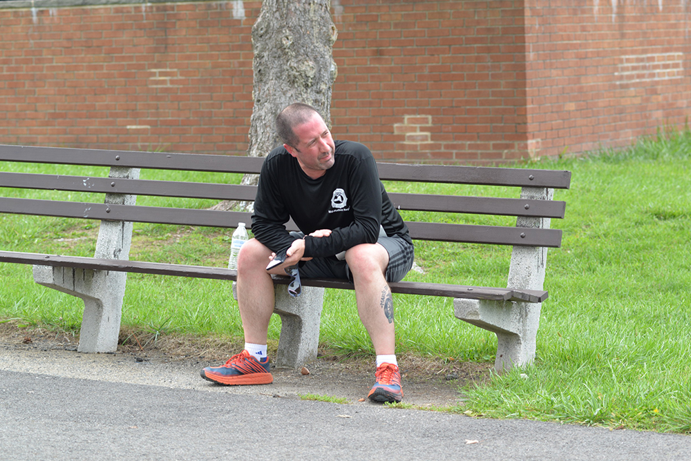 A man with a shaved head and a goatee sits on a wooden park bench. He is dressed in a black long-sleeve shirt and gray shorts, sporting red and blue running shoes. With a thoughtful expression, he looks off to the side. Grass and a brick wall are in the background, as if contemplating his next donation to charity.