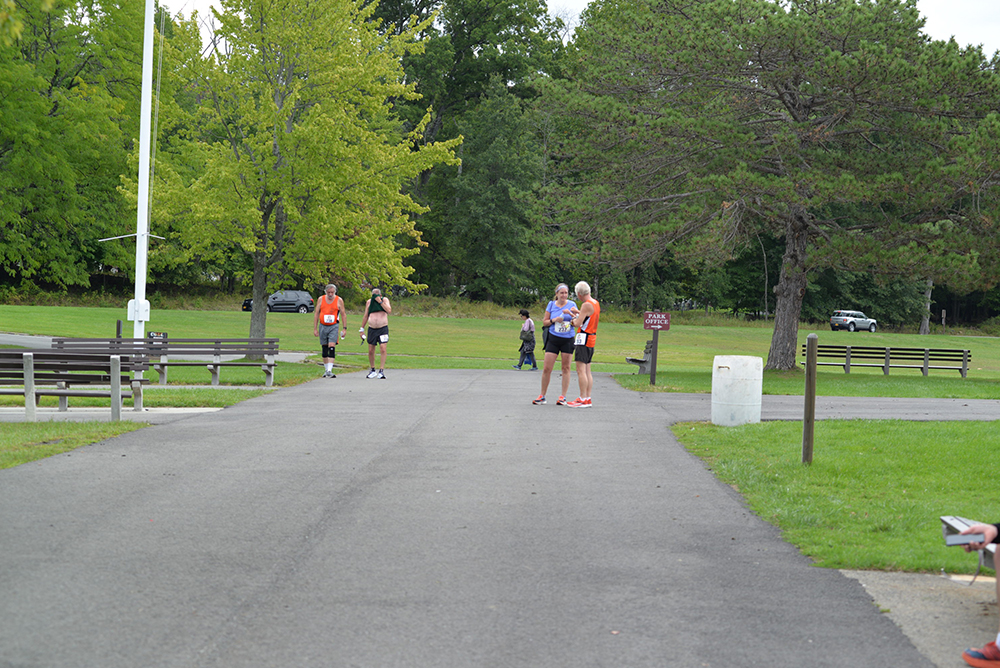 A group of people stand and talk on a paved pathway in a park, discussing an upcoming charity event. Several benches and trees with green foliage surround them. A person in running gear jogs in the background, while another walks. A car is partially visible among the trees, possibly there to collect donations.