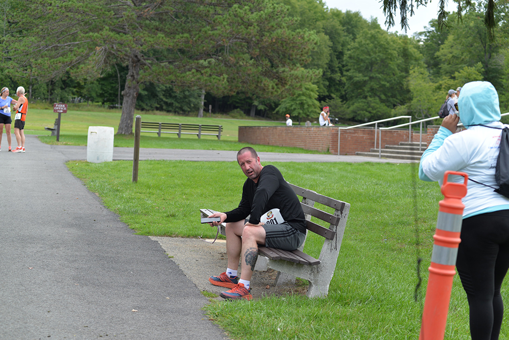 A man sits on a park bench, dressed in athletic gear. He holds a phone in one hand and a water bottle labeled "Donate" in the other. In the foreground, a person wearing a white top and dark pants appears to be gesturing or taking a photo. A few people are walking in the background.