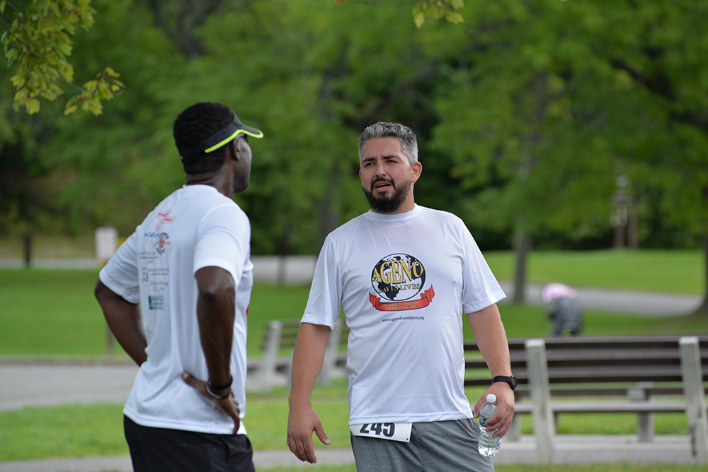 Two men wearing white athletic shirts are standing and talking outdoors in a park. One man has his hands on his hips, and the other holds a water bottle. Both appear to be participating in a charity race, as indicated by their race bib numbers. Green trees and a pathway are in the background.