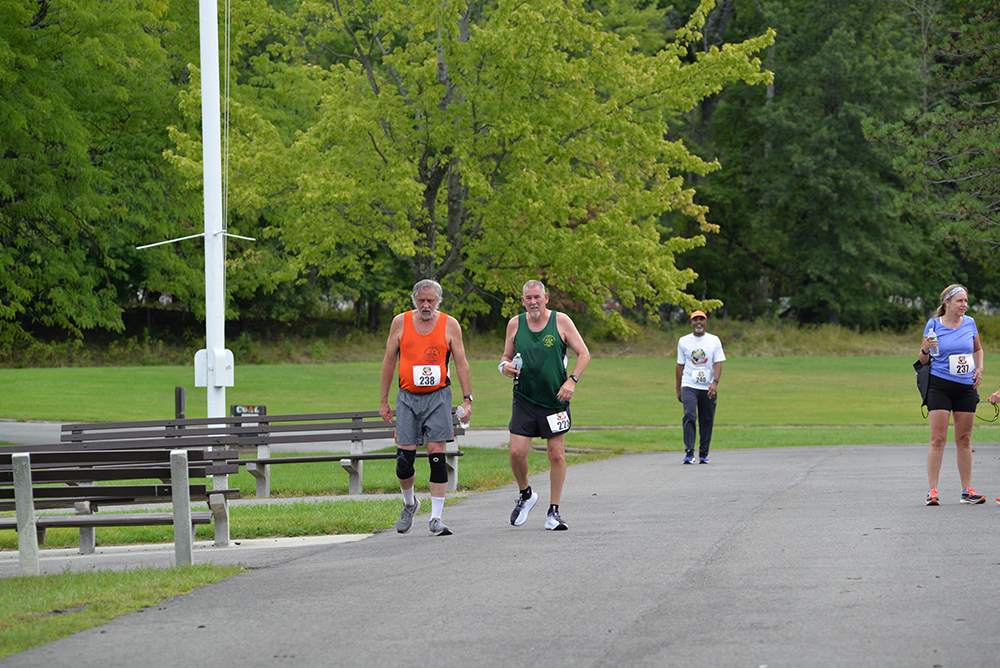 Two men with race bibs, one in an orange tank top and the other in a green tank top, run on a paved path in a park. Behind them, another runner and a person in casual clothes are visible. Another runner is to the side, all within a green, tree-filled landscape during a charity event to donate water.