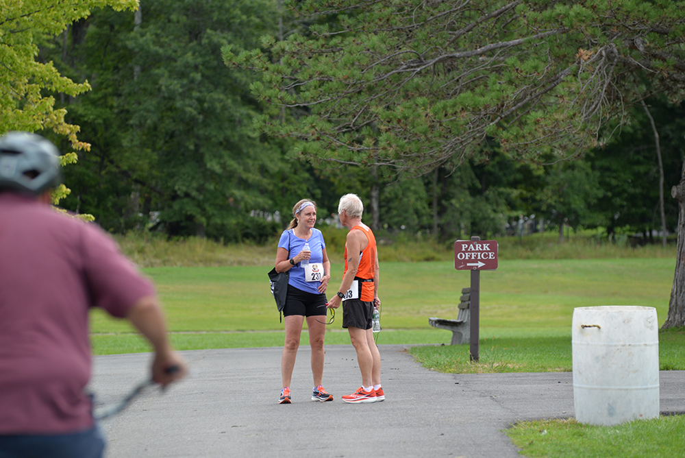 Two individuals are standing on a paved park pathway in athletic attire, conversing about the upcoming charity run. One is wearing a purple shirt and black shorts, the other an orange shirt and black shorts. A cyclist in a red shirt is seen in the foreground, near a "Park Office" sign.