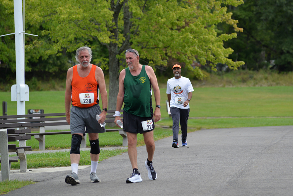 Three people are walking on a path in a park. The two in the foreground, participating in a charity race, are wearing race numbers—one in an orange tank top and the other in a black tank top. The person in the background is wearing a white t-shirt. Trees and a bench are also visible.