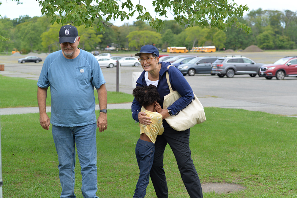 Two adults and a child are outdoors near a parking lot with school buses in the background. One adult, wearing a blue cap and glasses, is hugging the child. The other adult, in a blue shirt and baseball cap, stands beside them, smiling after making a generous donation to the local charity.