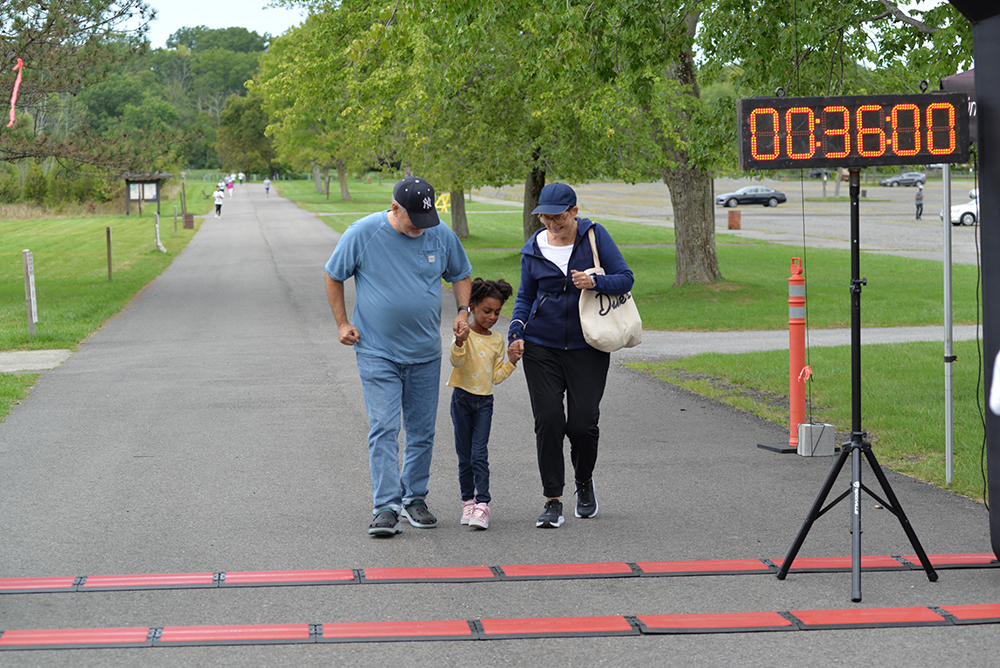 A man, woman, and young girl are walking together on a paved path towards a race finish line, which shows a time of 36 minutes. Surrounded by green trees and grass, with parked cars in the background, they are participating in a charity event to raise donations for those in need.