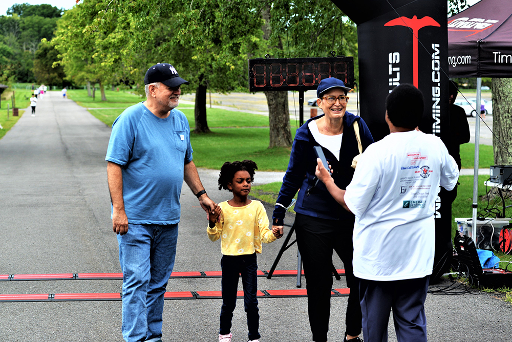 A man and woman, holding hands with a young girl, smile as they stand near a finish line at an outdoor charity event. The woman engages with another participant wearing a race bib. Trees and paths are visible in the background under an overcast sky, reminding everyone of the importance to donate for clean water initiatives.