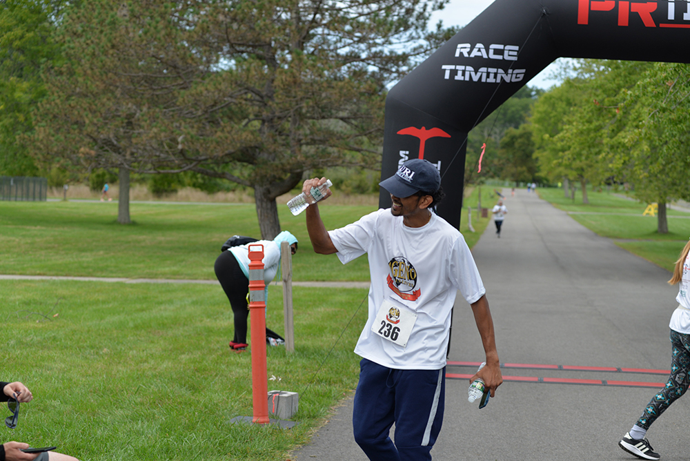 A runner wearing a white shirt, blue pants, and a cap is crossing the finish line at a charity race event, holding water bottles in both hands. A few other participants and onlookers are visible in the background, with a grassy area and trees surrounding the path.