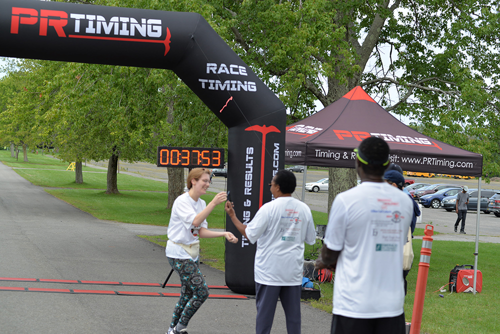 Runners in white shirts cross the finish line of a race under a black PR Timing arch. A digital clock displays a time of 00:37:53. A red and black PR Timing tent is set up nearby with people standing around it, donating to charity. Trees and parked cars are visible in the background.