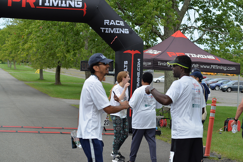 Participants in a race wearing white shirts celebrate and congratulate each other near the finish line. Behind them, an inflatable race arch displays "Race Timing" signage next to a tent with more people, timing equipment, and a website URL for donations to charity. Trees line the background.
