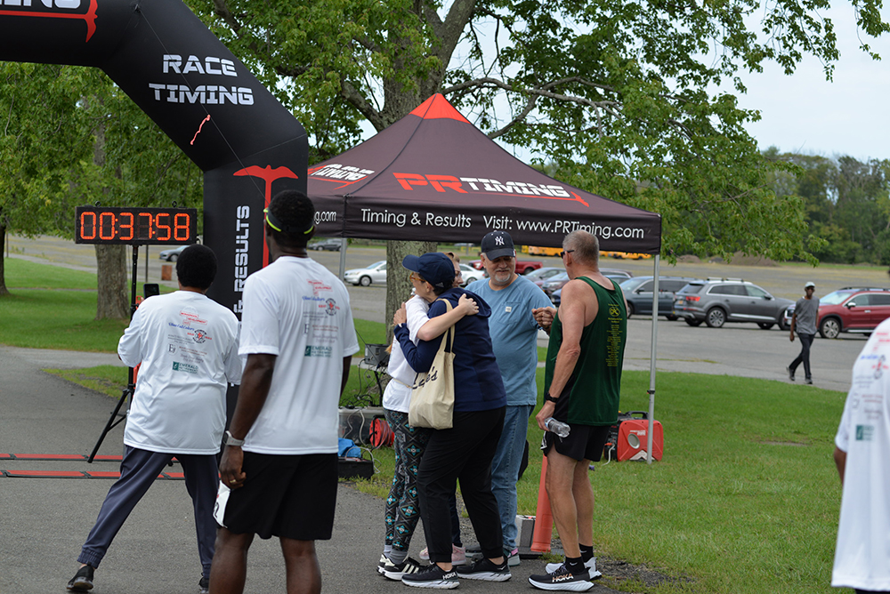 A group of people stands near a race finish line and a timing tent with "PR Timing" on it. A digital clock displays 03:37:58. Some participants wear running gear; one person is being embraced by someone else. Trees and parked cars are visible in the background, emphasizing the event's charity atmosphere.