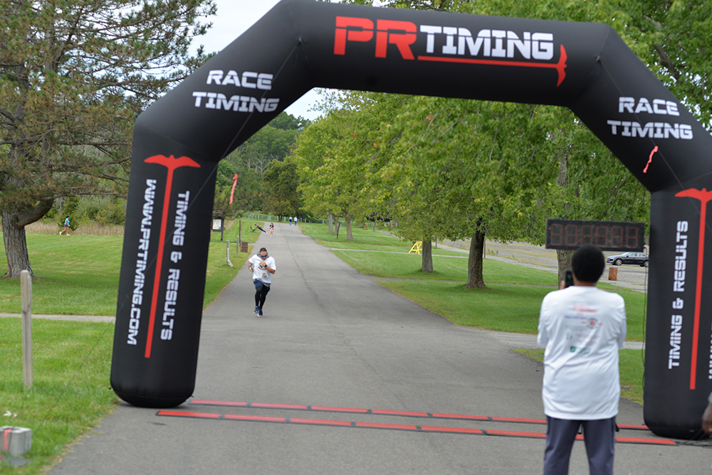 A person is running towards the finish line of a race, marked by an inflatable arch that reads "PR Timing." Another individual stands near the finish line, possibly overseeing the timing. The event, which supports charity, is taking place in a park with green trees lining the path.