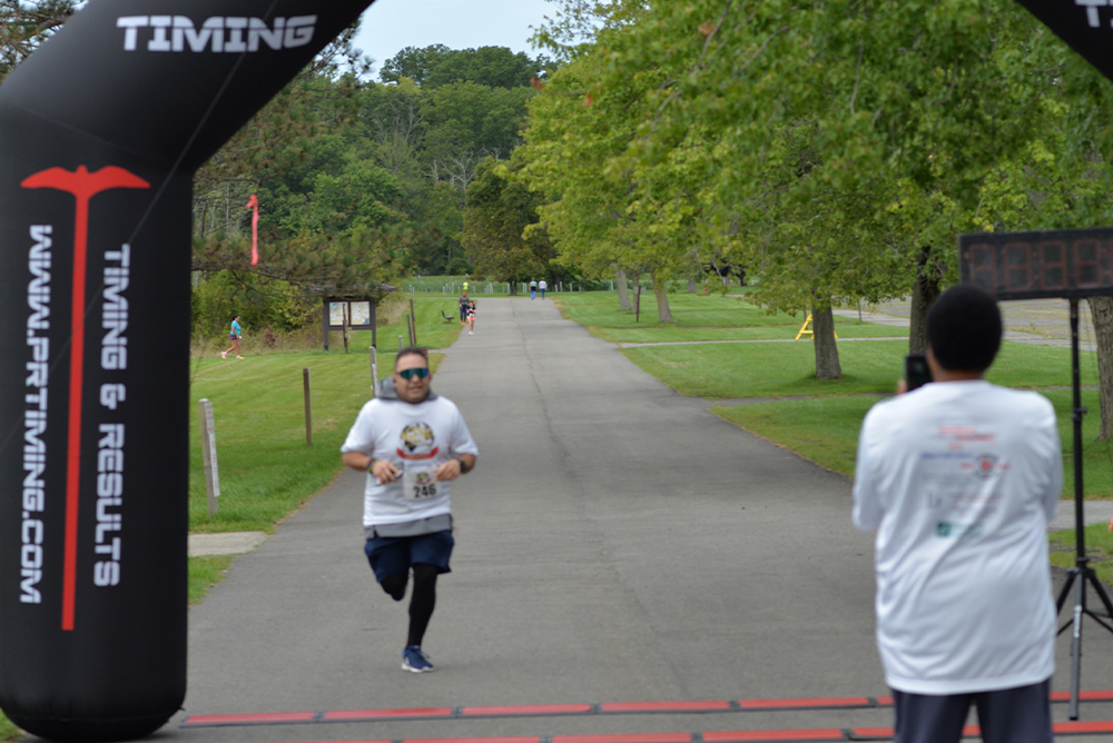 A runner wearing sunglasses and a white shirt is approaching the finish line of a race. The race banner reads "TIMING" and "www.pttiming.com." Another person in a white shirt stands nearby, possibly capturing the moment for charity. Green trees line the background.