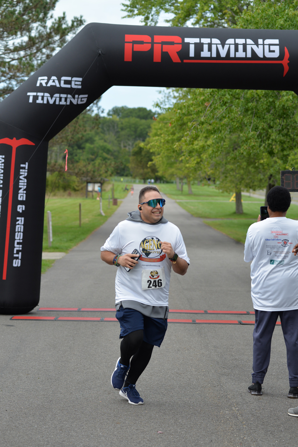 A man wearing sunglasses, a white shirt with the number 246, and dark pants runs under a black archway labeled "PR TIMING." Another person stands to the side, encouraging participants to donate. Trees and a grassy field are visible in the background.