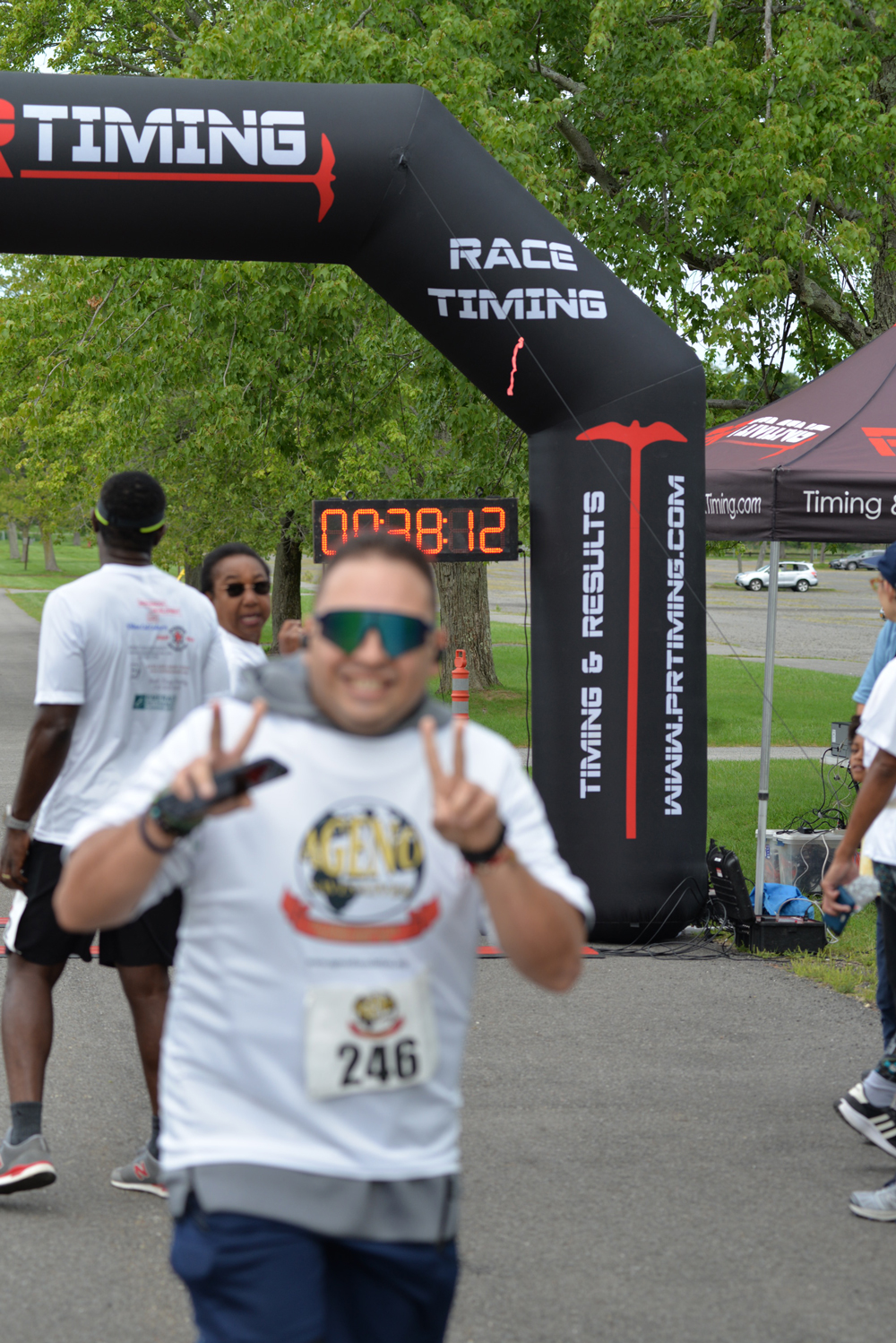 2nd-annual-5k-living-water-run-img50 A runner wearing a white shirt and sunglasses flashes a peace sign as he passes under a black inflatable arch with a digital timer reading "00:38:12." Other participants and event staff are seen in the background. The arch displays the text "Race Timing," celebrating an event dedicated to charity.
