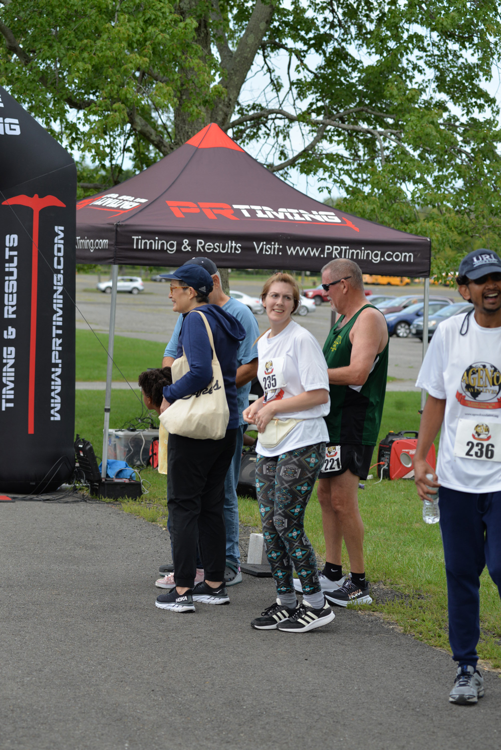 A group of runners stand near a timing and results tent with the logo "PR Timing" at an outdoor event. Some are smiling and chatting, while others seem to be preparing for or resting after the run. Nearby, a table offers water bottles for those who wish to donate to charity. A few cars are parked in the background.