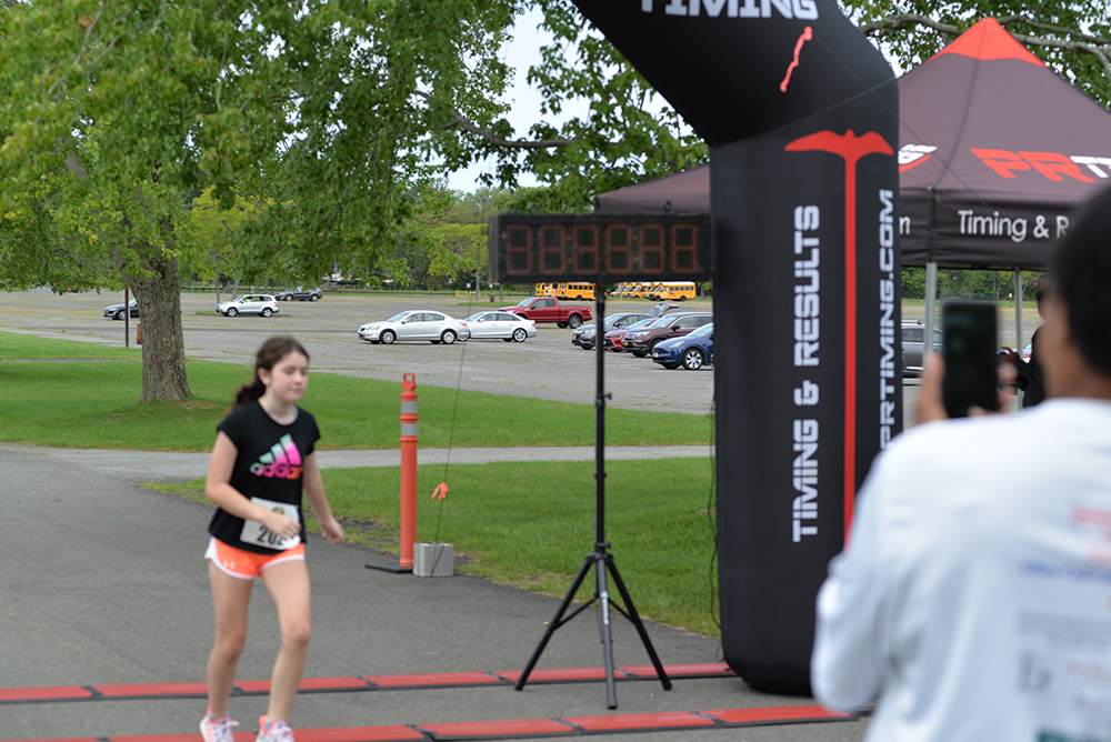 A runner wearing a black and pink outfit crosses a finish line under an inflatable arch. An individual in the foreground is taking a photo. The background includes parked cars, trees, and grassy areas. A timing display and a tent with "Timing & Results" are visible at the charity event.