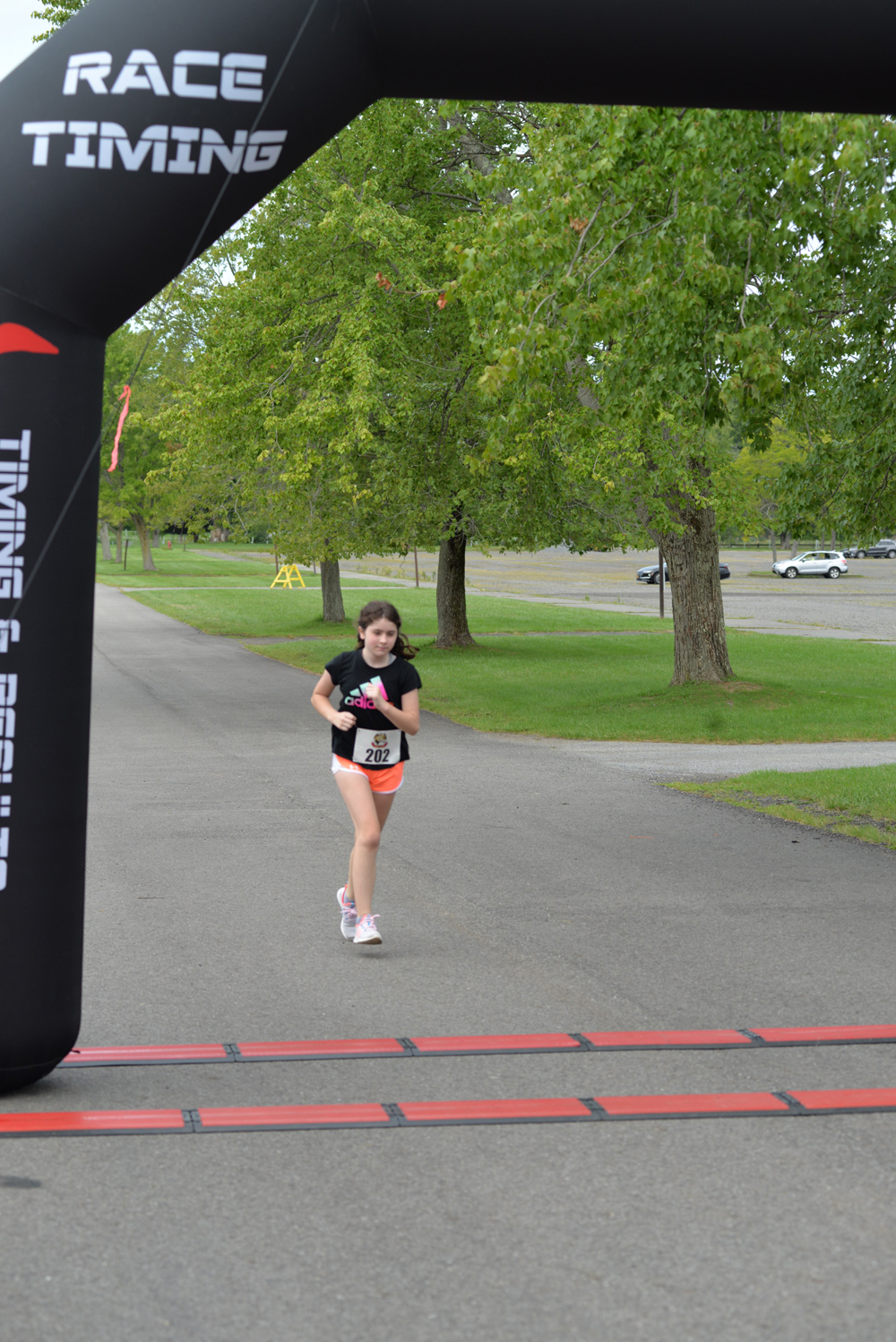 A young girl wearing a black shirt and white shorts is running towards the finish line of a charity race. She is holding a pink water bottle, and there are trees and parked cars in the background. The finish line arch is black and has "RACE TIMING" written on it, urging everyone to donate generously.