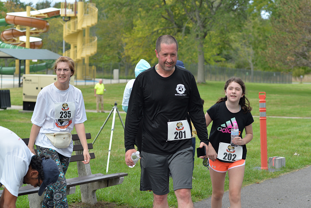 A group of people participate in a charity race at a park. A man wearing a black shirt and holding a water bottle walks next to a young girl in a black shirt and pink shorts. A woman in a white shirt rests near a bench. Bib numbers 201, 202, and 235 are visible, encouraging donations for the cause.