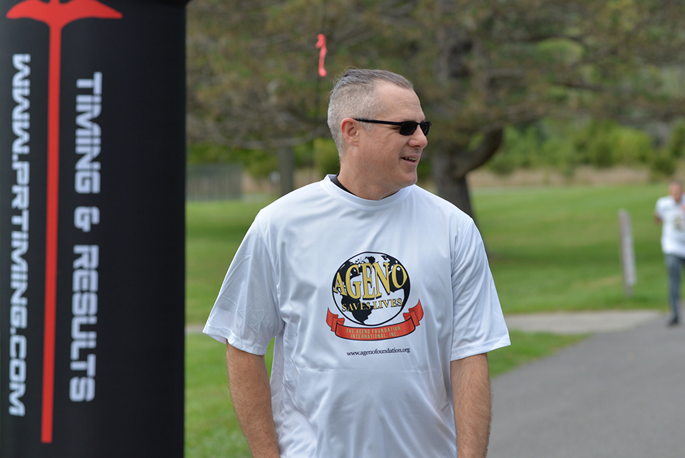 A man wearing sunglasses and a white event t-shirt stands outdoors, next to a sign that reads "PR TIMING Timings & Results." The background, with green trees and a path, hints at a charity event aimed at raising donations for clean water initiatives.