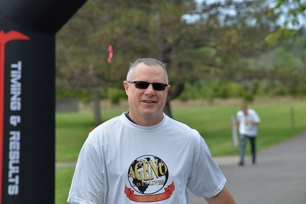 A man wearing sunglasses and a white "A Geno Saves Lives" t-shirt smiles while walking outside. He is under an inflatable arch with "TIMING & RESULTS" printed on the side. Another person in a similar t-shirt can be seen in the background, supporting the charity event amidst trees and grass.