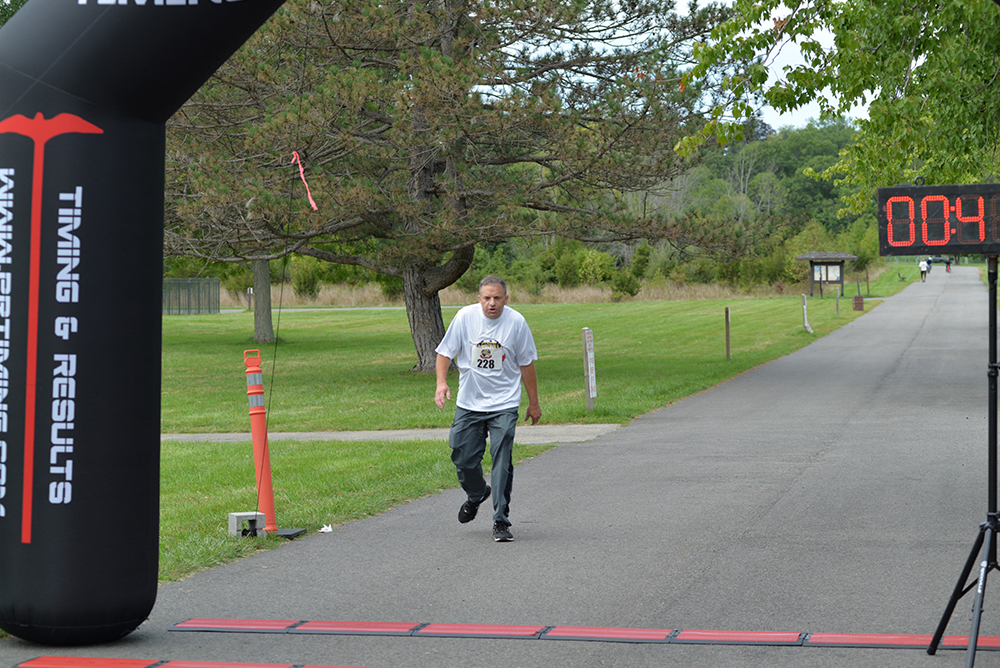 A runner, wearing a white shirt with number 28, approaches the finish line of a race. The digital timer shows 004. The path is surrounded by greenery with trees in the background, and a black arch displaying "Timing & Results" frames the finish area. Nearby, volunteers offer water to runners.