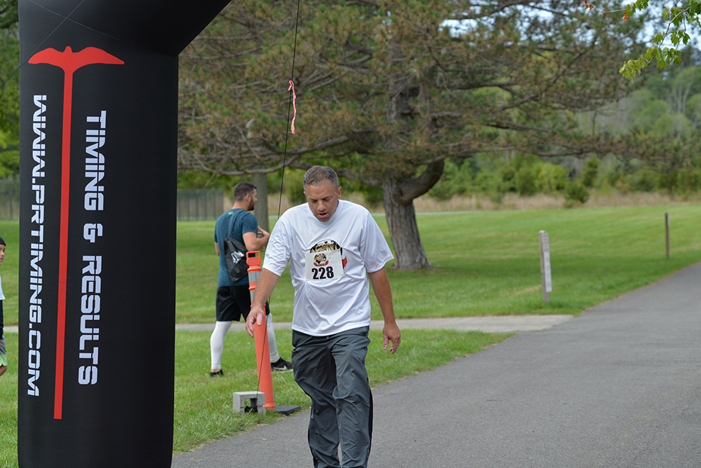 A man wearing a white shirt and grey pants is reaching the finish line of a race. He appears tired and is catching his breath. The finish line banner displays "Timing & Results" ahead of lush, green trees bordering the pathway, with water stations reminding participants to donate to charity causes.