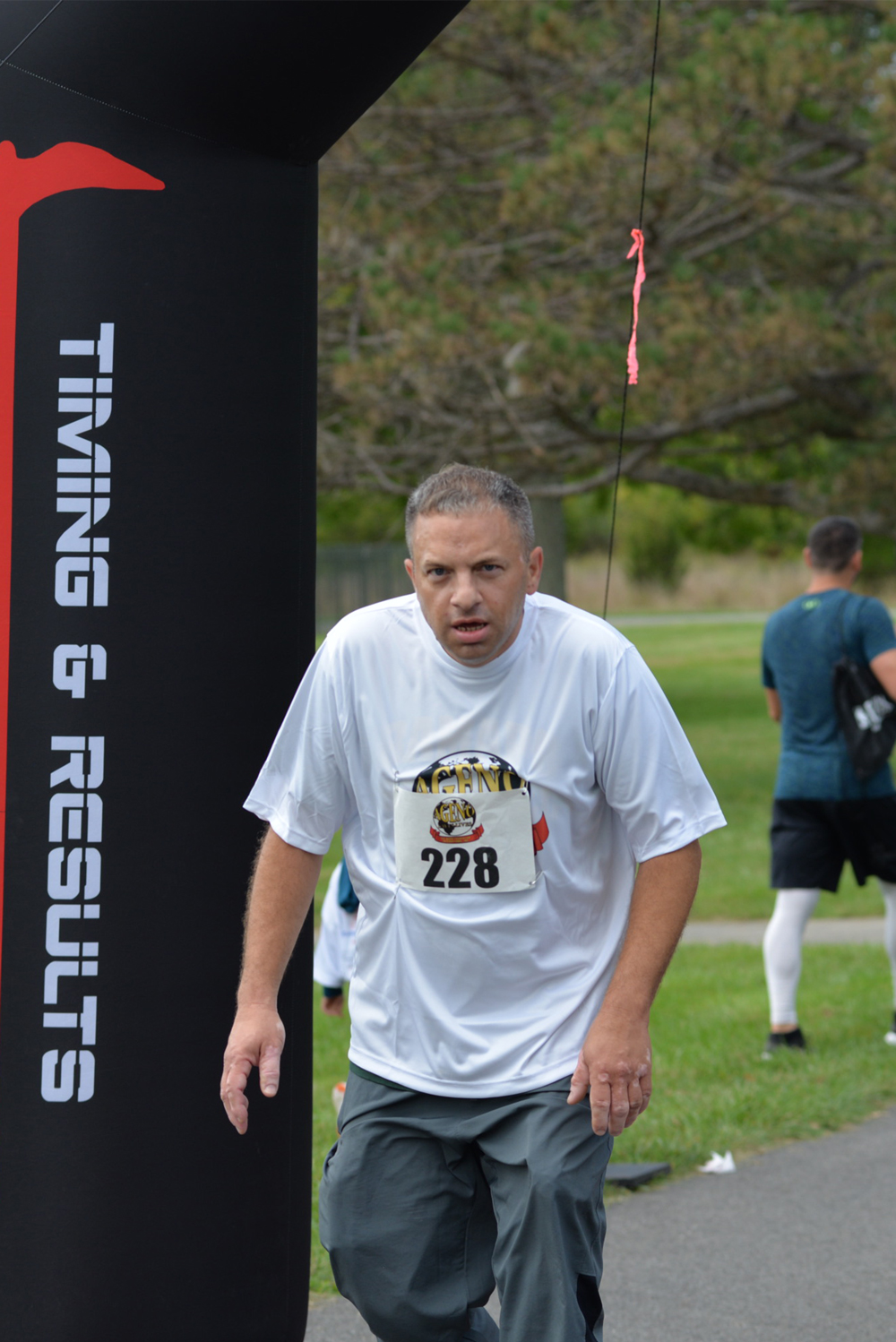 A man wearing a white shirt and gray shorts with the race number 228 is crossing a finish line. A black banner with the text "TIMING & RESULTS" is visible on the left. The background includes other runners and a park setting with trees and grass, highlighting the charity event where donations were encouraged.