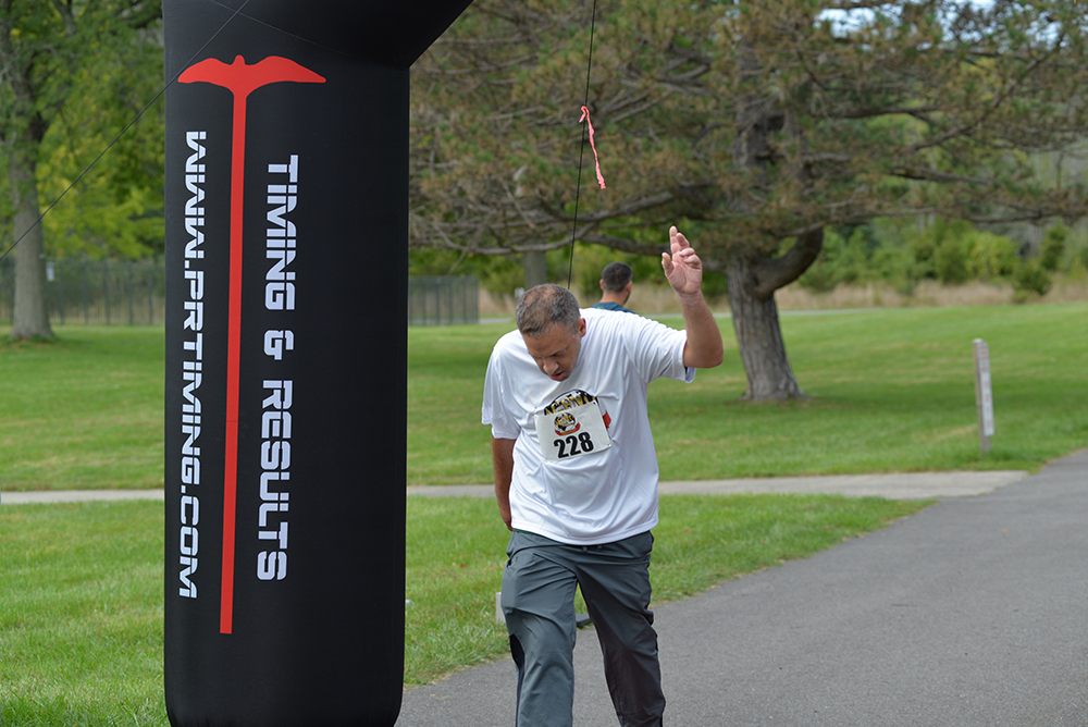 A man wearing a white t-shirt and gray pants crosses the finish line in a race, marked by an archway labeled "Timing & Results." He raises one arm in the air, celebrating his victory. The background includes greenery and another person walking, holding out a bottle of water as if making a refreshing donation.
