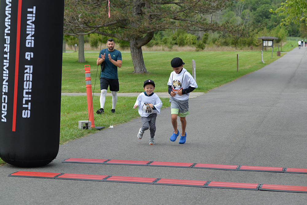 Two young boys wearing race bibs and white shirts run towards the finish line of a charity race, marked by a black and red inflatable arch. An adult claps and cheers them on from the side, while volunteers offer water in the background against the lush greenery of the park.