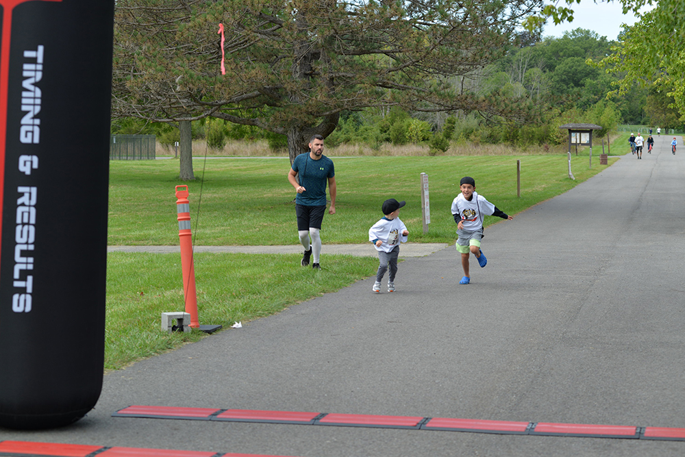 A man and two young boys are running on a paved path in a park toward a finish line. The boys are ahead of the man and focused on the finish. The black inflatable arch with "TIMING & RESULTS" stands at the end, marking not just their victory but also their commitment to charity. Trees and greenery form the backdrop.