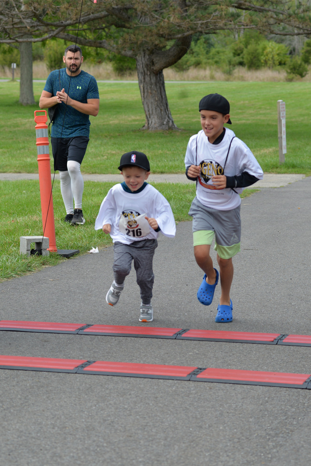 Two young boys are running across the finish line of a race, smiling and looking happy. The younger boy is in front, wearing a baseball cap and a race bib numbered 216. An adult jogger in a teal shirt watches them from the background on a green park trail, holding a bottle of water to donate to the runners.