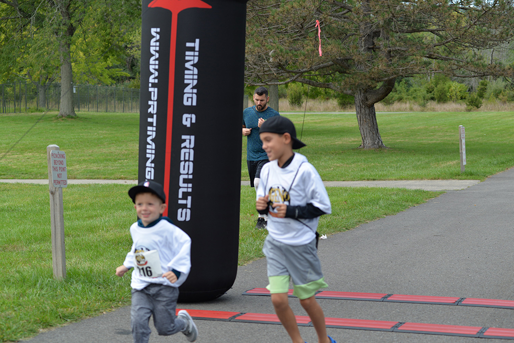Two children with number bibs are running a race, crossing a finish line with a black inflatable arch labeled "TIMING & RESULTS." A man in the background is walking on the path. The outdoor setting features grass and trees, and water stations nearby encourage donations for charity.