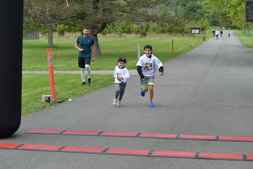 Two boys, wearing event t-shirts, run across a finish line on a paved path in a park, with a man jogging and other participants further in the background. The charity fun run set amidst lush green trees and grass aims to raise funds for clean water initiatives.