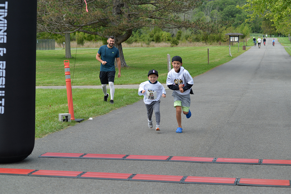 Two young boys in athletic wear and numbered bibs run toward the finish line of a charity race on a paved path. An adult jogs behind them, smiling. The setting is a park with green grass, trees, and other people in the distance. A large inflatable marker is on the left.