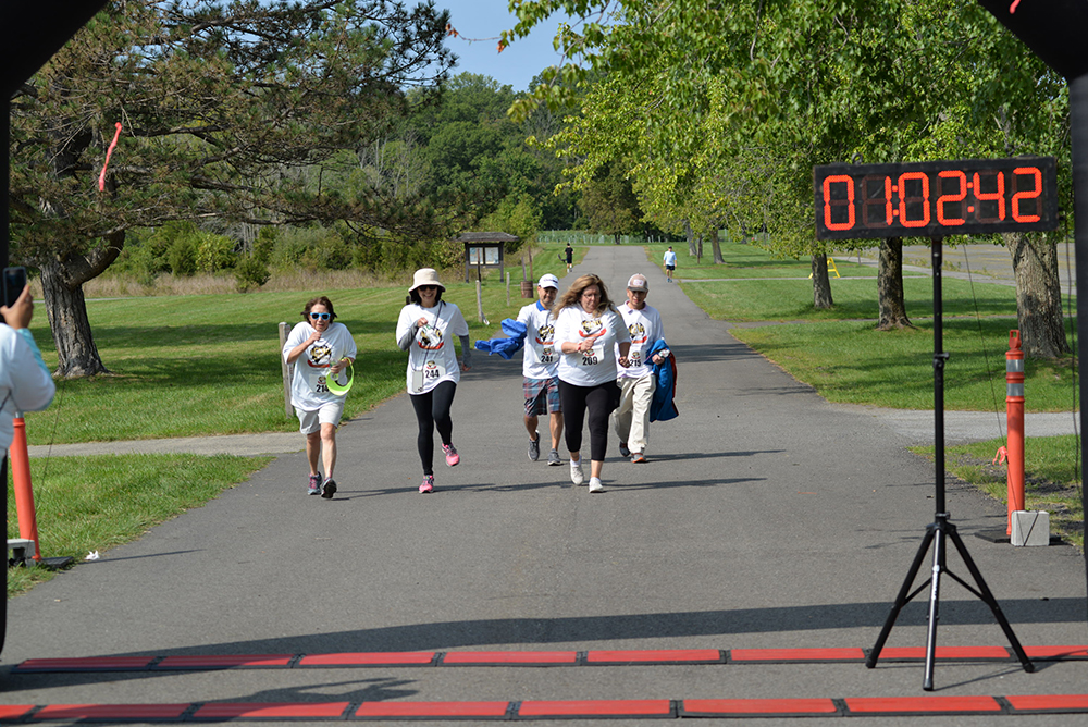 Five people wearing white T-shirts with logos run towards a finish line in a park, as a digital timer shows 1 hour, 2 minutes, and 42 seconds. Trees and greenery form the backdrop while an individual captures the moment with a camera. This charity run event encourages participants to donate for clean water initiatives.