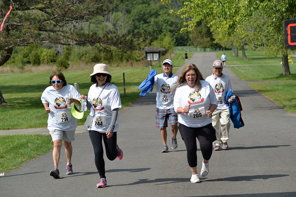 Five people wearing matching white t-shirts with numbers jog down a paved park path, smiling and holding small trophies. They seem to be participating in a fun run event to raise money through donations. Trees and grass are visible in the background.