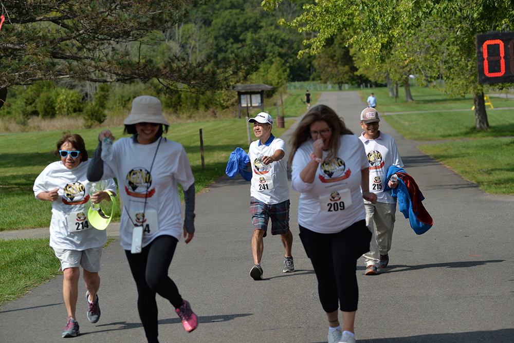 A group of five people, wearing white t-shirts with matching designs, are walking or jogging on a paved path in a park. Some are holding jackets and sipping water. The path is surrounded by greenery, and a large digital timer displaying "8" is visible in the background as part of their charity event to encourage donations.