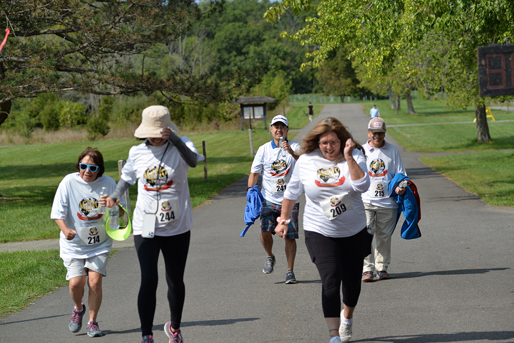 A group of people wearing white event t-shirts with race numbers, walking on a paved path in a park. They look tired but determined as they participate in the charity event. The background includes green grass, trees, and a wooden shelter. It is a sunny day.