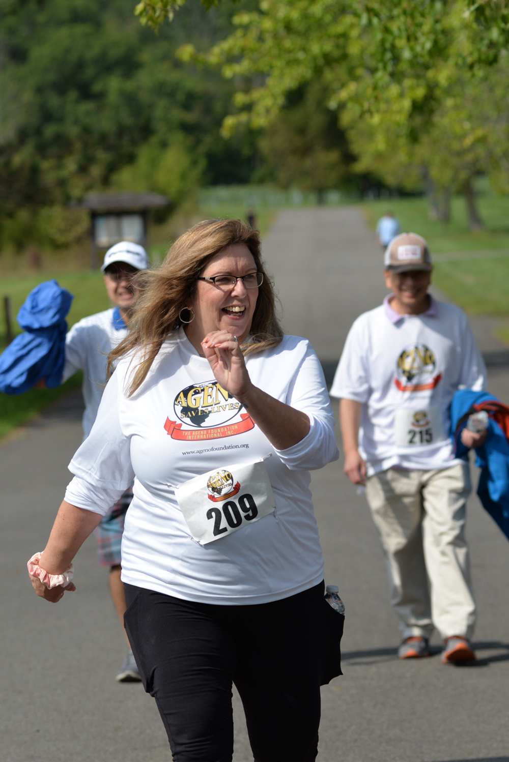 A woman in glasses and a white shirt with the "Agent in the Zone" logo runs on a paved path wearing race bib number 209. Two others follow her; one, with a thumb up, has race bib 215. Trees and green grass line the path on a sunny day, as runners support this charity event.